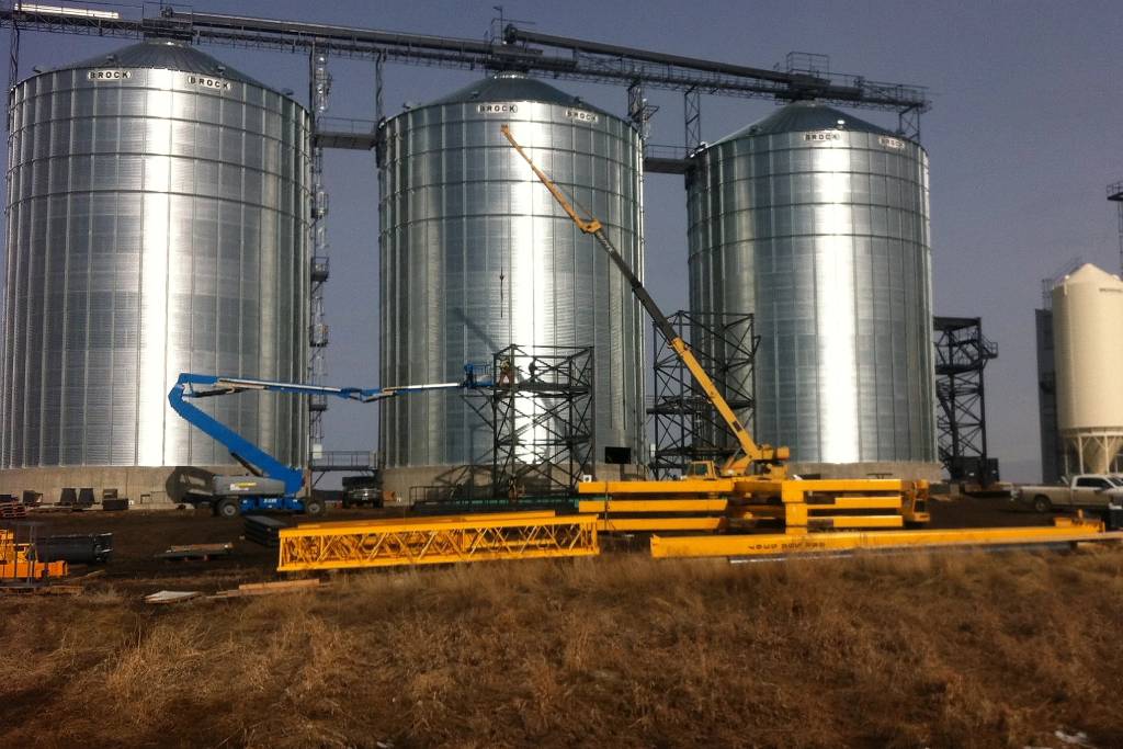 Three steel grain silos. Cranes work in the foreground to construct the buildings.